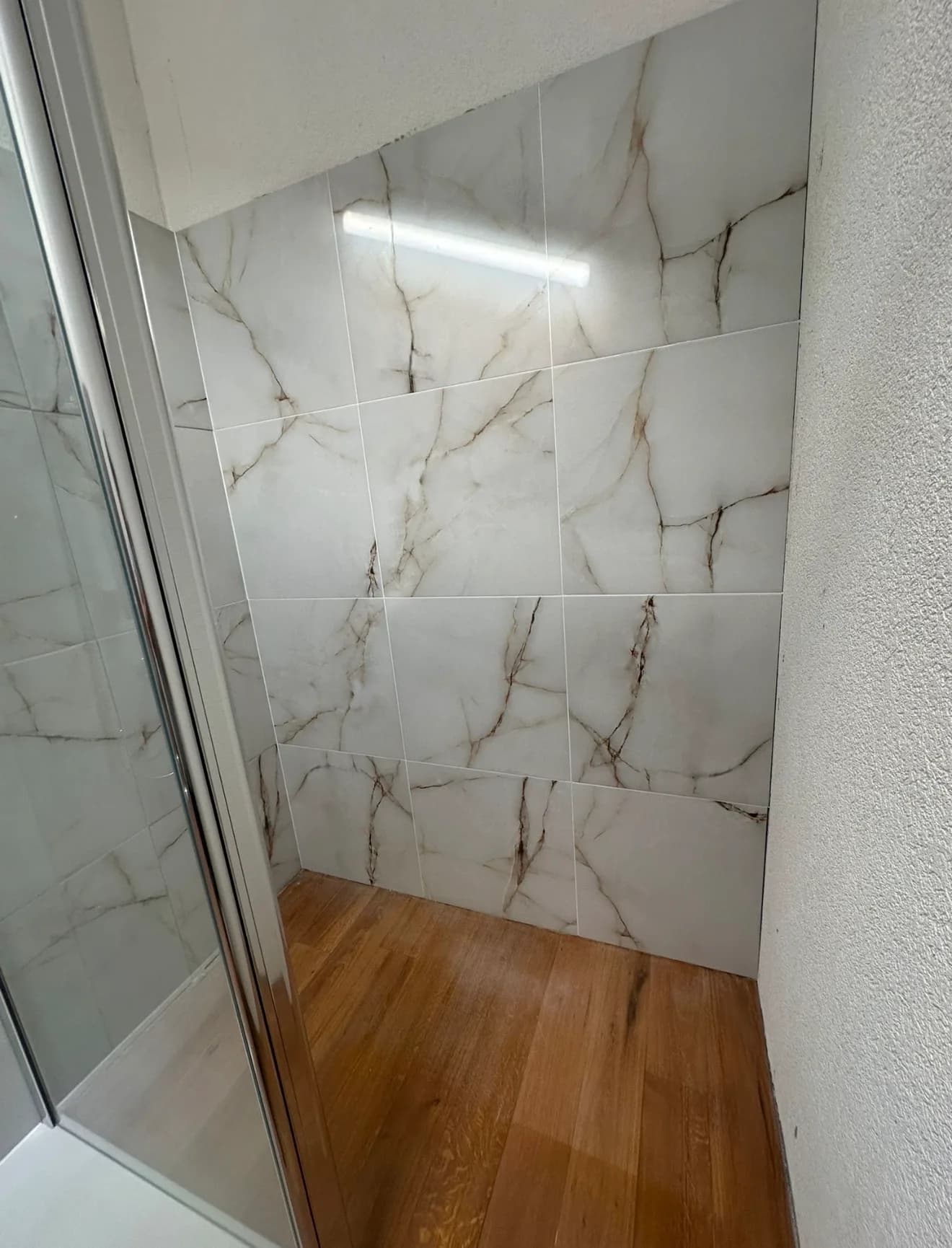 Alcôve sous un plafond en pente, mur en carreaux blancs veinés effet marbre et sol en lames d'aspect bois, à côté d'une paroi de douche vitrée