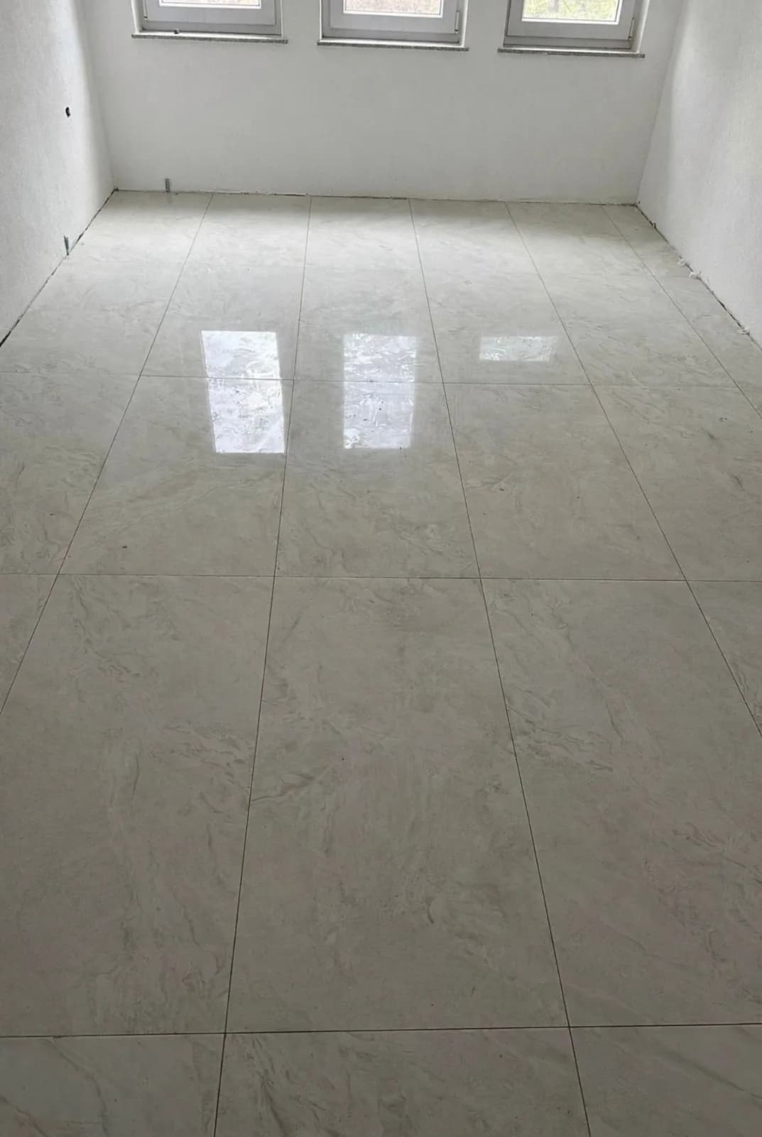 Pièce vide au sol entièrement couvert de grands carreaux gris clair effet marbre, où se reflètent les fenêtres