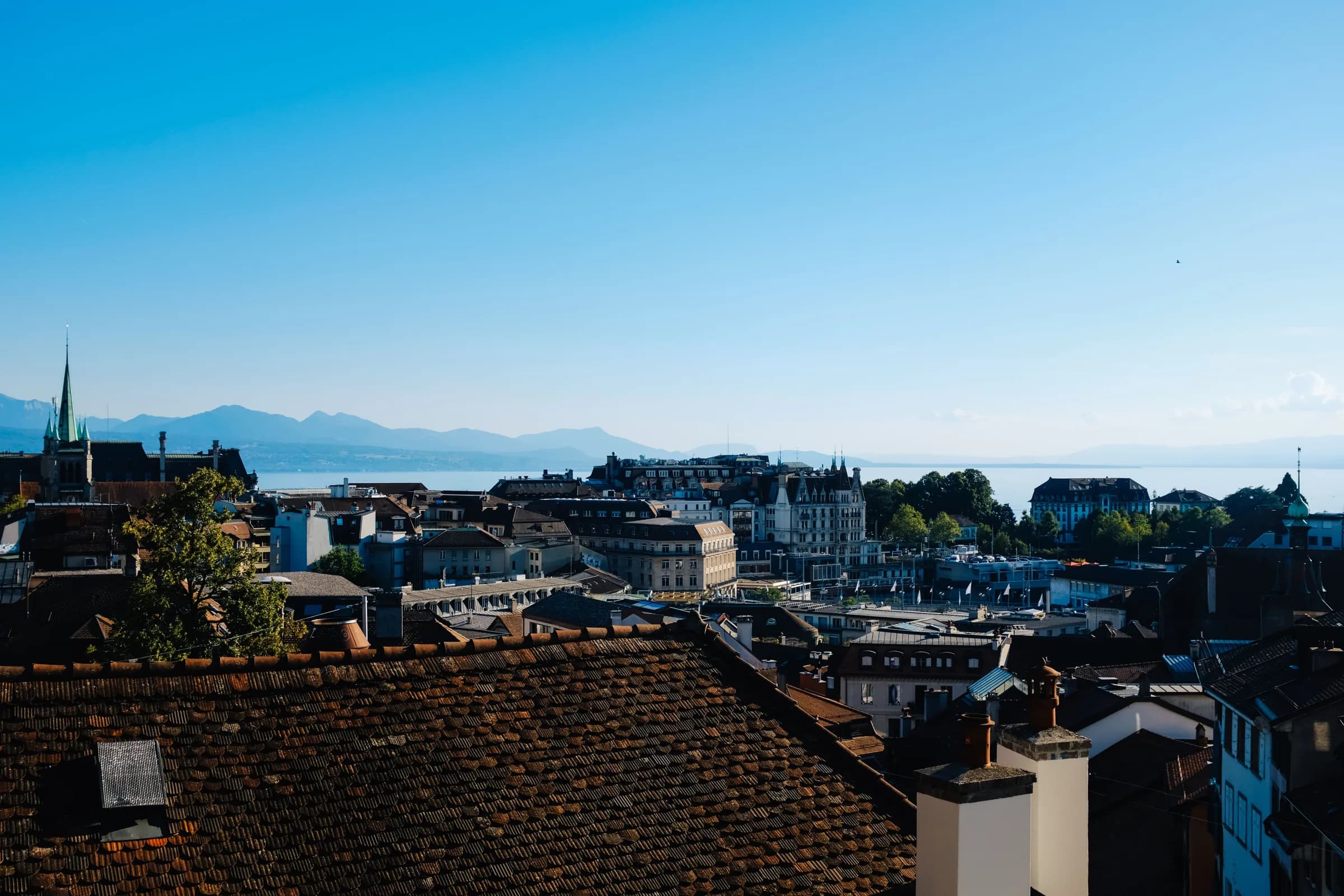 Les toits de Lausanne vus d'en haut par temps clair, un clocher à flèche verte, puis le lac Léman et les montagnes sous un grand ciel bleu