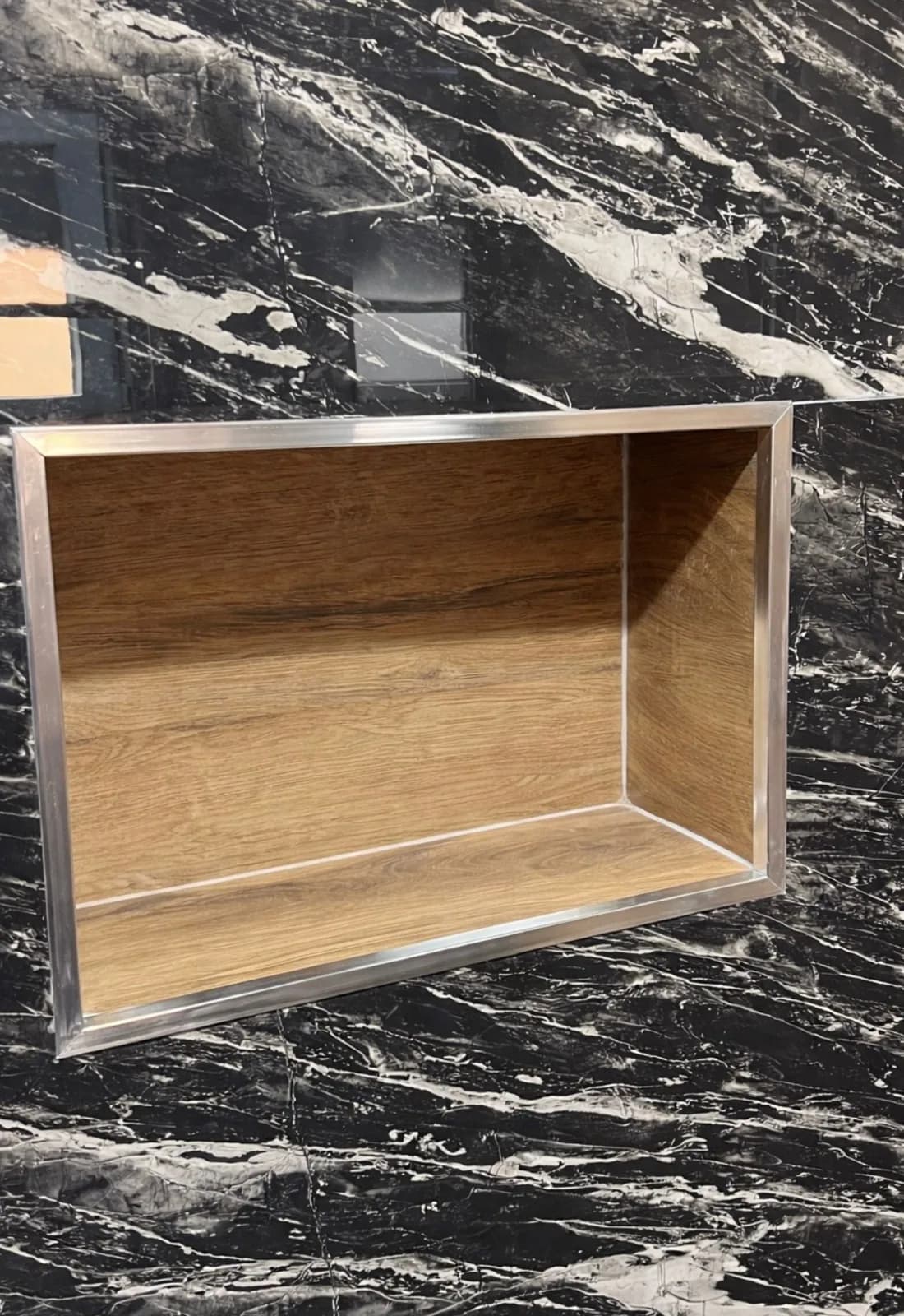 Niche murale encastrée, fond en carreaux d'aspect bois et cadre métallique brossé, dans un mur en carreaux noirs veinés de blanc effet marbre