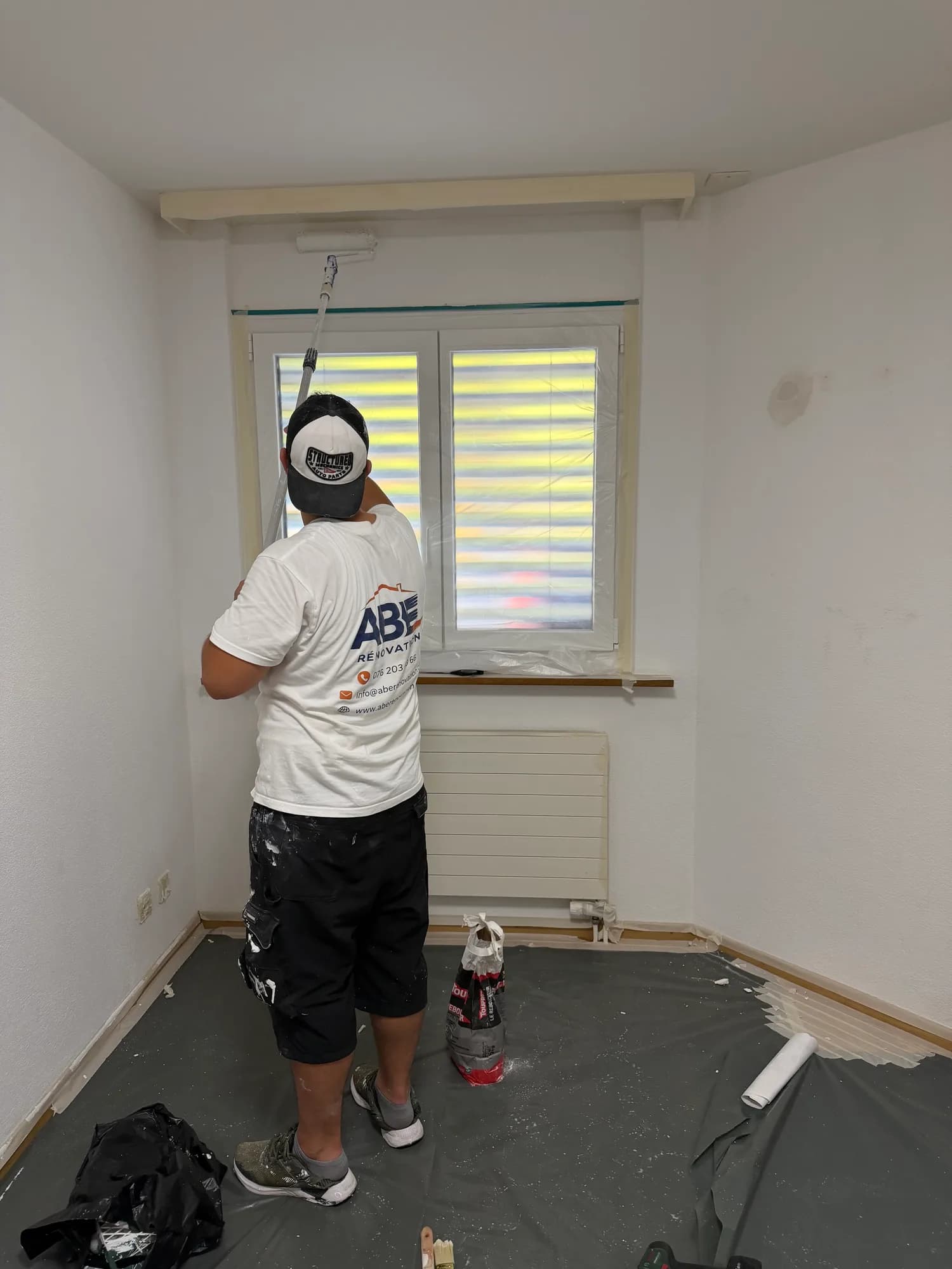 Un ouvrier de dos, t-shirt au logo ABE Rénovation, peint au rouleau le haut d'un mur au-dessus d'une fenêtre protégée par une bâche