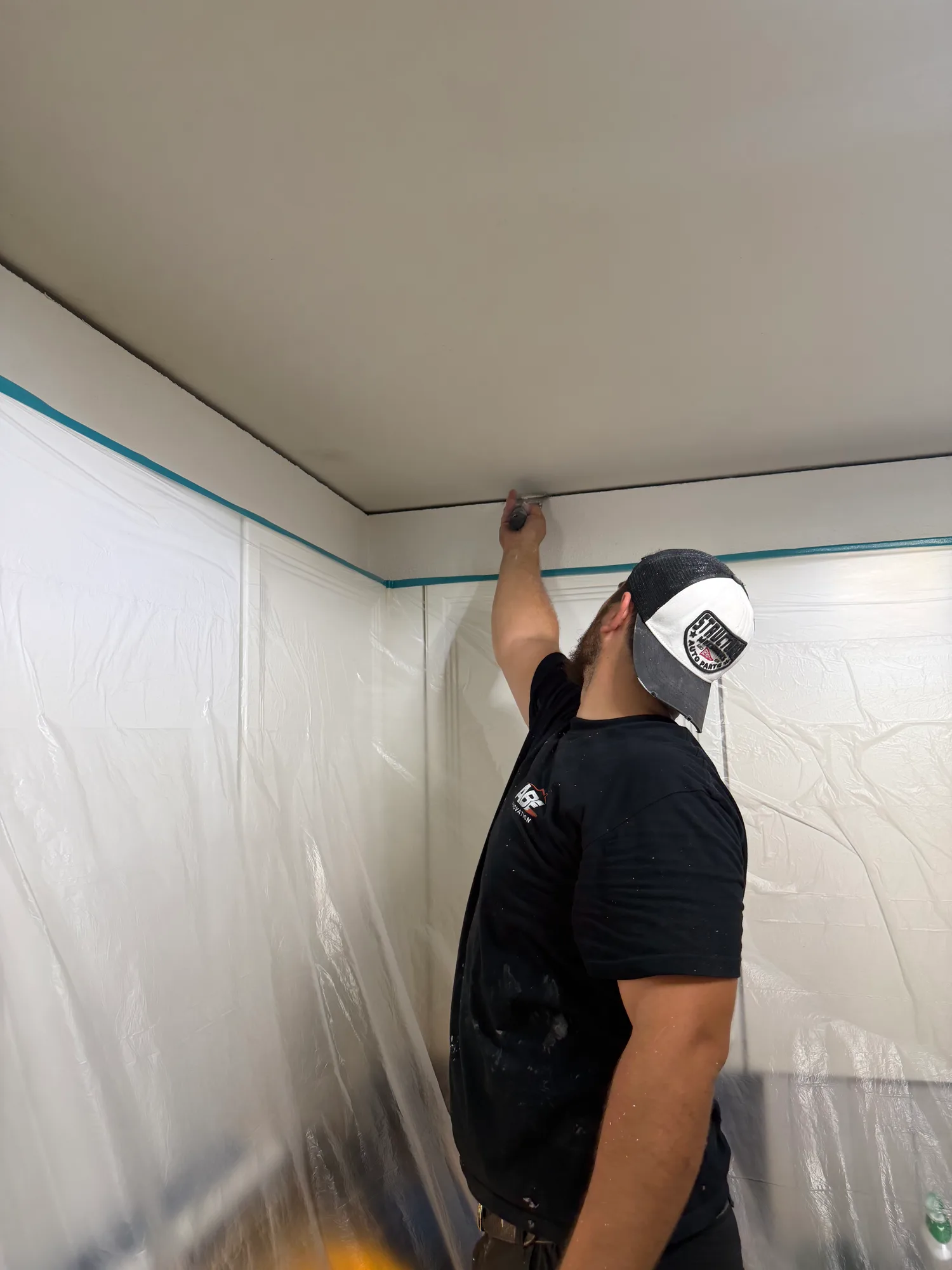 Un ouvrier au t-shirt ABE Rénovation peint au pinceau la jonction entre le mur et le plafond, les murs protégés par une bâche et un ruban de masquage bleu
