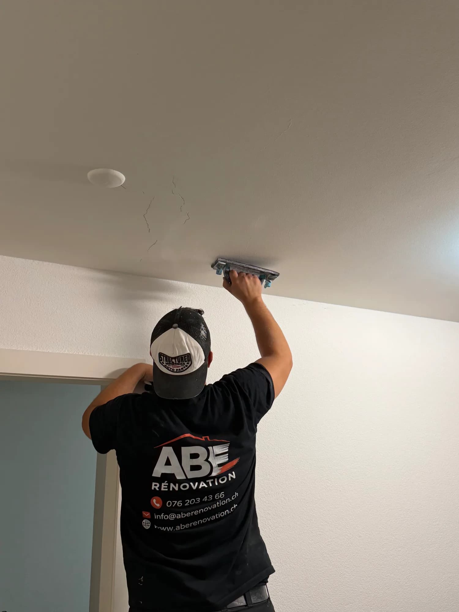 Un ouvrier de dos, t-shirt au logo ABE Rénovation, ponce à la main un plafond blanc qui porte des fissures