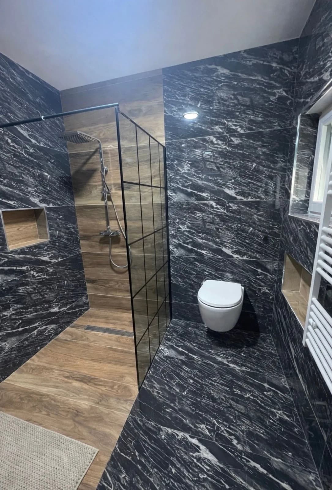 Salle de bains en grands carreaux noirs veinés effet marbre : douche à l'italienne derrière une paroi vitrée, niches encastrées et WC suspendu