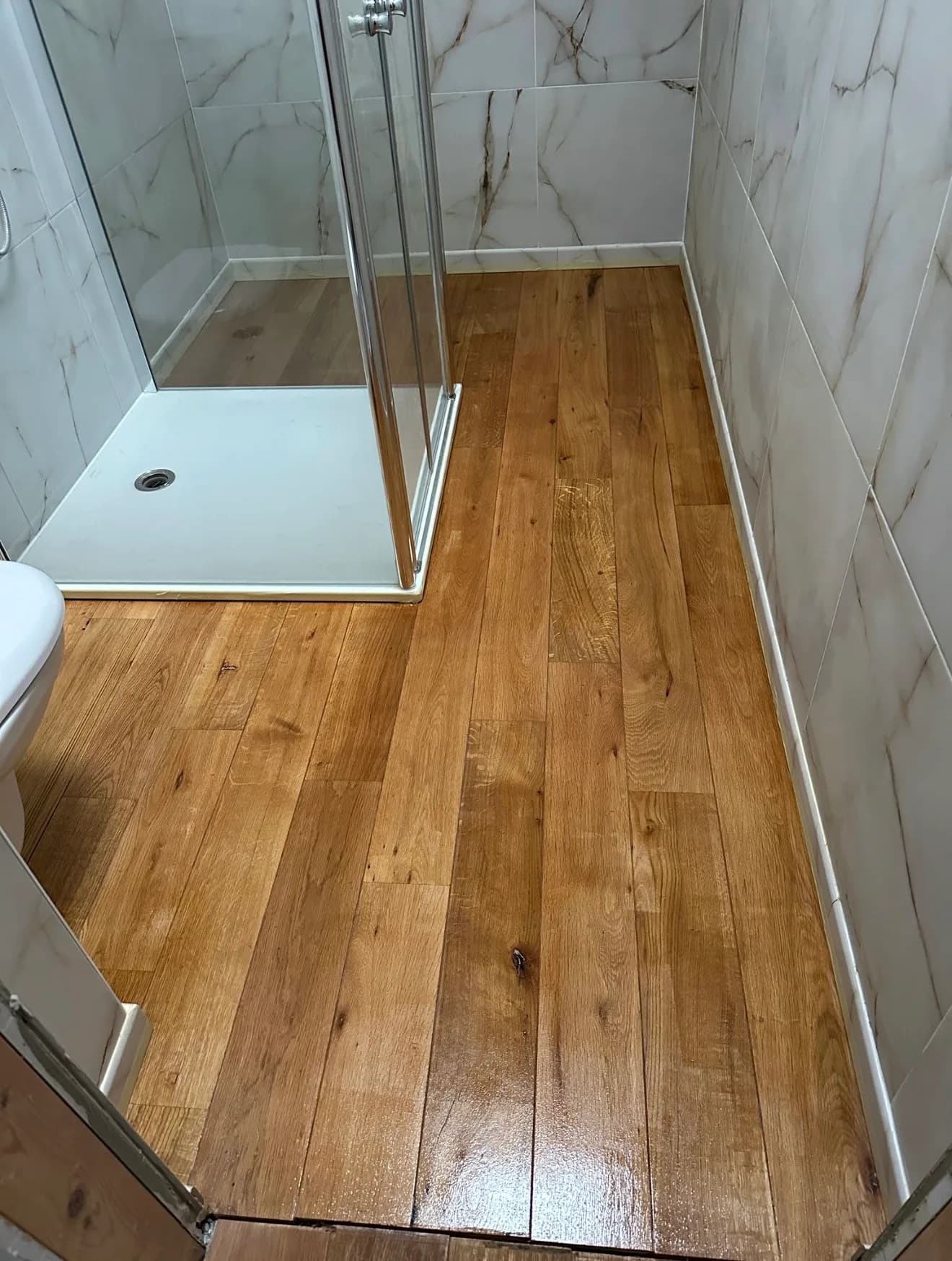 La même salle d'eau terminée : sol en lames d'aspect bois teinte miel et brillant, murs en carreaux blancs veinés effet marbre, cabine de douche vitrée