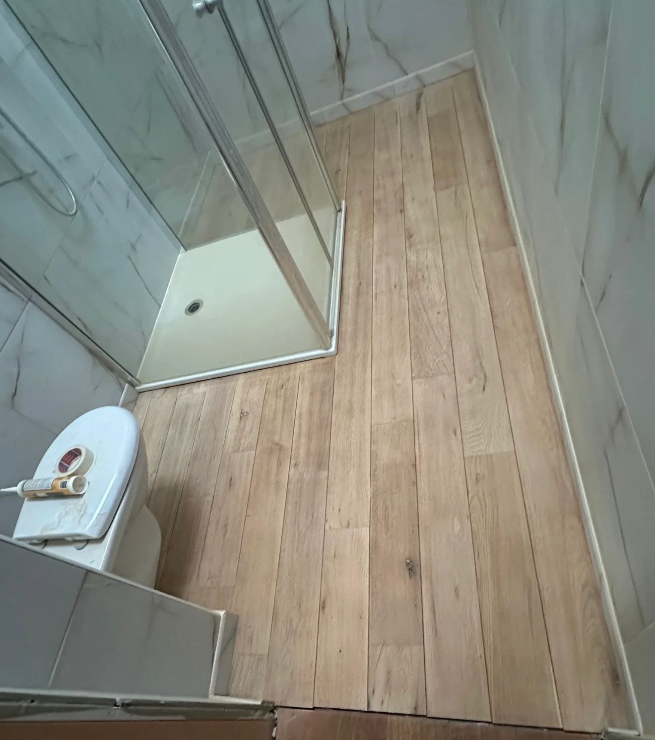 La salle d'eau pendant les travaux, vue d'en haut : sol en lames d'aspect bois clair et mat, cabine de douche vitrée, un rouleau de ruban adhésif et une cartouche de mastic posés sur le WC